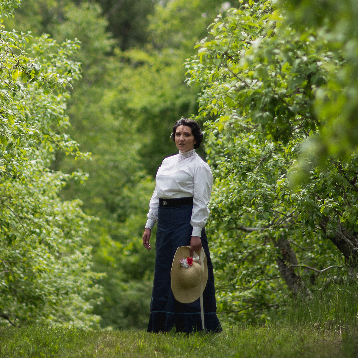 Woman in white and navy turn of the centry dress standing in an orchard surrounded by green trees