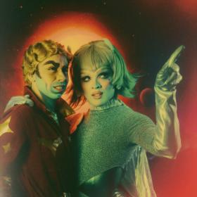 Briar and Rusty in intergalactic garb stand against an otherworldly sky. Briar points offscreen.