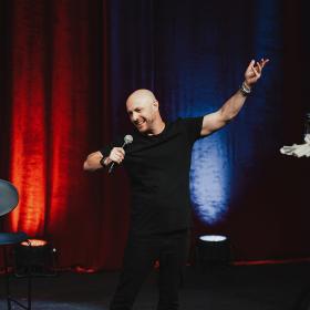 Comedian Avi Nussbaum holds a microphone with his opposite arm outstretched