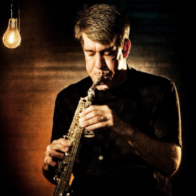 Daniel Ian Smith playing a soprano sax with eyes closed while being lit by a single hanging lightbulb
