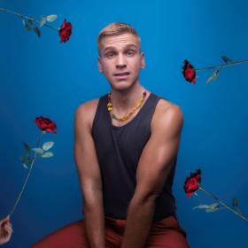 Michael John Ciszewski faces the camera with a quizzical expression with 4 red roses around him against a blue background