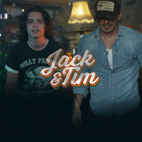 Father and son duo Jack and Tim face the camera in a dimly lit room. They wear casual clothing and Tim has a baseball cap on obscuring his face.