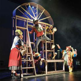 actors climb around on a set like a window made of scaffolding wearing absurd and colorful costumes