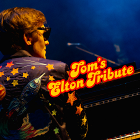 Performer Tom Cridland as Elton John sits at a piano facing away from the viewer