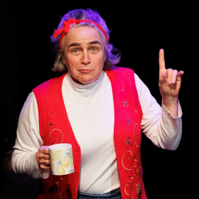 Performer Cindy Pierce wears a red vest over a white shirt and points one finger in the air while looking perplexed and holding a coffee cup